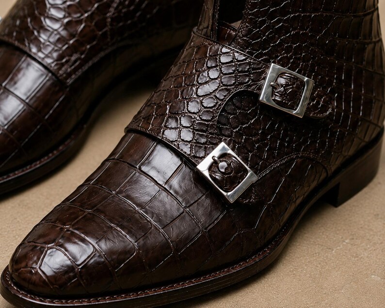 May include: Close-up of a pair of brown leather boots with a crocodile skin texture. The boots feature silver buckles with a skull design. The leather has a glossy finish, and the boots appear to be new.