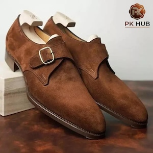 Könnte beinhalten: Braune Wildleder-Mönchschuhe mit Schnallenverschluss. Die Schuhe haben eine spitze Zehenpartie und einen niedrigen Absatz. Sie werden auf einem weißen Block und einer braunen Oberfläche präsentiert. Das Logo "PK HUB" ist oben rechts sichtbar.