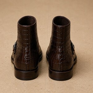 May include: A pair of brown leather ankle boots with a crocodile texture. Each boot has a buckle closure on the side and a stacked heel. The boots are displayed on a neutral-toned surface.