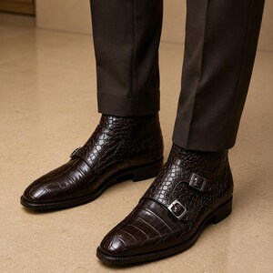 May include: A pair of dark brown leather ankle boots with a crocodile texture. The boots feature double silver buckles and are worn with dark brown trousers. The boots have a pointed toe and a low heel.