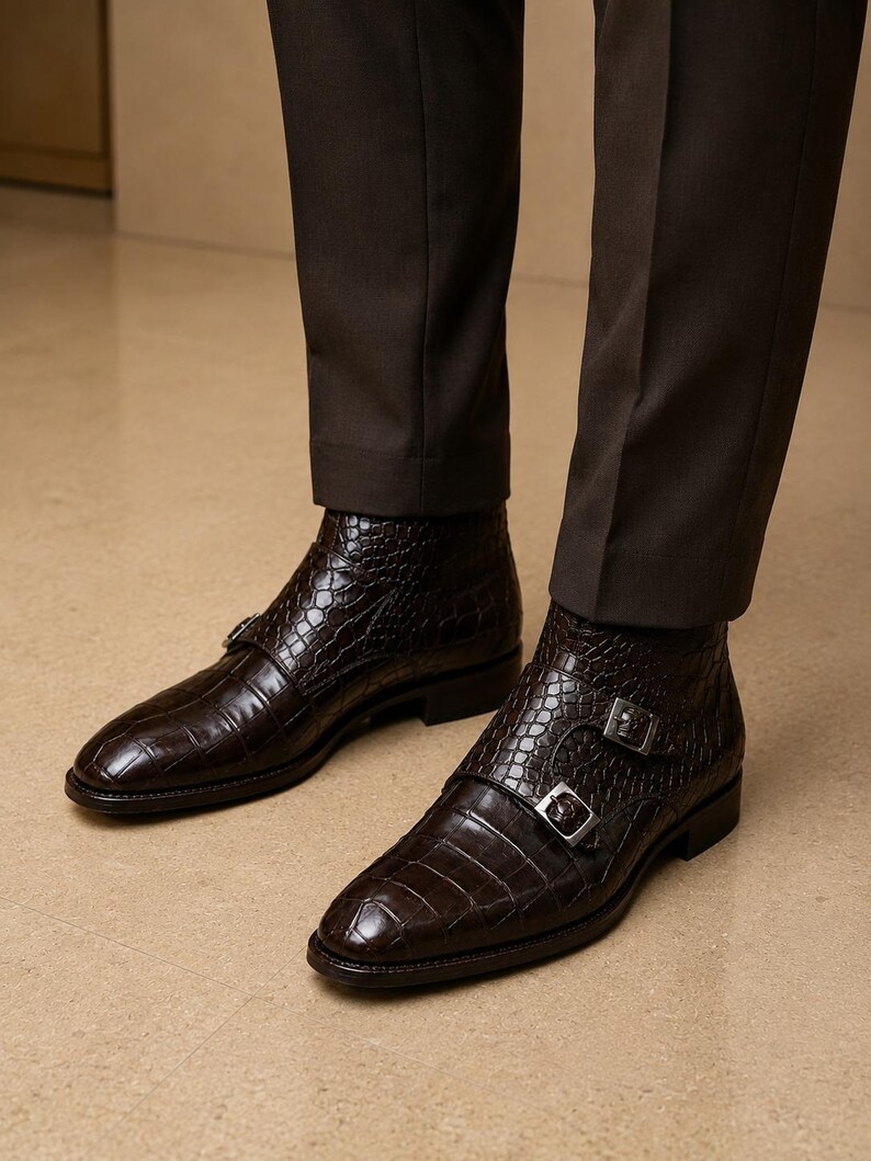 May include: A pair of dark brown leather ankle boots with a crocodile texture. The boots feature double silver buckles and are worn with dark brown trousers. The boots have a pointed toe and a low heel.