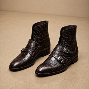 May include: A pair of brown leather ankle boots with a crocodile texture. The boots have a pointed toe, a double buckle closure, and a short shaft. The boots are displayed on a neutral background.