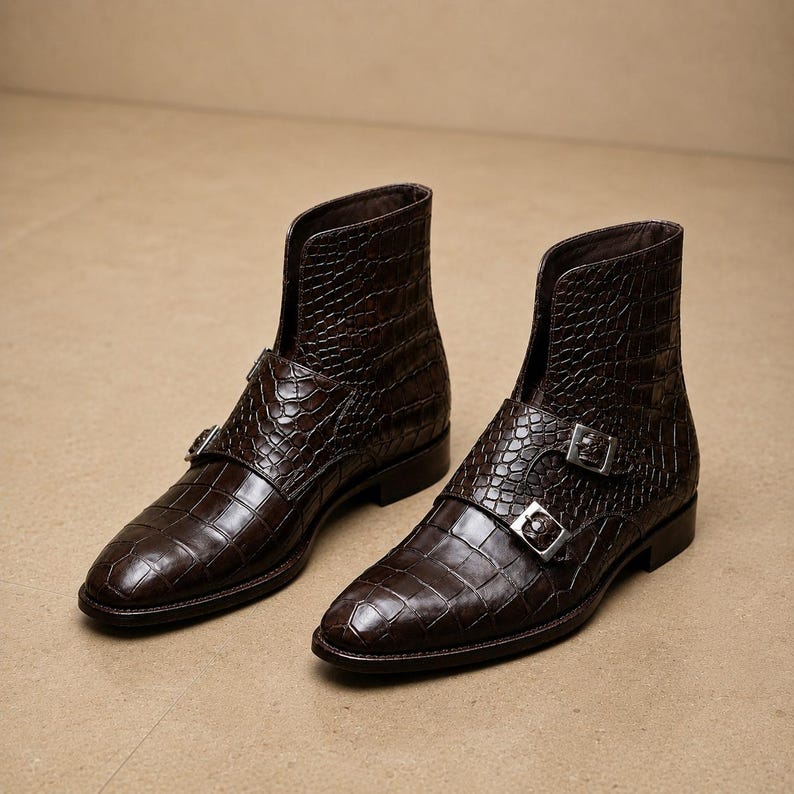 May include: A pair of brown leather ankle boots with a crocodile texture. The boots have a pointed toe, a double buckle closure, and a short shaft. The boots are displayed on a neutral background.