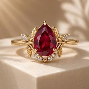 May include: A gold ring featuring a central pear-shaped red gemstone. The setting is adorned with metallic leaf motifs and small round white stones arranged at the top and bottom of the main gem, creating a detailed botanical-inspired design.