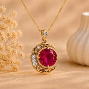 May include: A gold necklace featuring a crescent moon pendant with intricate filigree detailing. The pendant is set with a large, round, deep red gemstone in the center and accented with several small, clear, round-cut white stones.