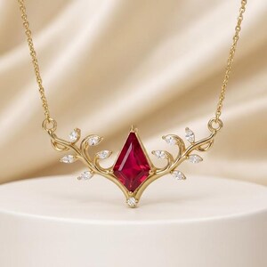 May include: A gold necklace featuring a central kite-shaped red gemstone flanked by two symmetrical vine-like branches. Each branch is adorned with small, clear marquise-cut stones, and a single small round clear stone sits at the base of the red gem.