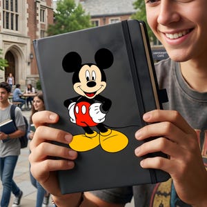 May include: A black notebook featuring a full-body image of Mickey Mouse in his classic red shorts, yellow shoes, and white gloves. The notebook has a black elastic band closure and the text "UNIVERSITY CAFE" is visible on the side.