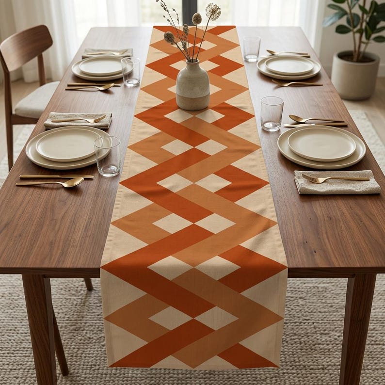 Peut inclure: Un long chemin de table rectangulaire pr&eacute;sentant un motif g&eacute;om&eacute;trique de bandes en zigzag entrelac&eacute;es dans des tons orange br&ucirc;l&eacute;, terre cuite et cr&egrave;me. Le chemin est centr&eacute; sur une table &agrave; manger en bois avec des assiettes blanches.