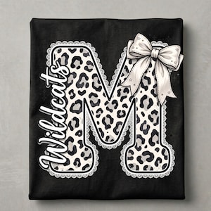 May include: A black t-shirt featuring a large letter M with a white and grey leopard print pattern. The letter is outlined in white lace trim and adorned with a large white bow. The word Wildcats is written vertically in white script.