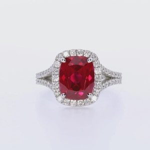 May include: A stunning ring with a large, cushion-cut, deep red gemstone, encircled by a halo of smaller, sparkling white stones. The split shank band is also embellished with white stones, creating a brilliant display of light and colour.