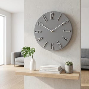 May include: A gray wall clock with white numerals and hands hangs on a light gray wall. A wooden shelf below holds a white vase with a green leaf, a stack of books, and a small potted succulent. A gray sofa is visible in the background.