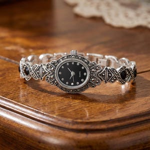 Op de afbeelding: Zilverkleurig quartz horloge met een zwarte wijzerplaat en decoratieve versieringen. Het horloge heeft een reeks schakels met zwarte accenten en kleine, heldere stenen rond de wijzerplaat. Het horloge ligt op een houten oppervlak.