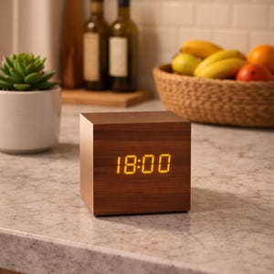 Puede incluir: Un reloj digital cúbico de madera que muestra las 18:00 en un tono amarillo-naranja cálido. El reloj está sobre una encimera, con una suculenta en una maceta blanca a la izquierda y una cesta de frutas en el fondo.