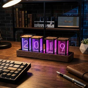 Puede incluir: Un reloj digital con marco de madera que muestra el número "2019" en dígitos morados brillantes. El reloj está sobre una base de madera. Un teclado y un bolígrafo están en el escritorio. El fondo incluye una estantería con libros y un altavoz Marshall.