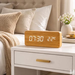 Puede incluir: Un reloj digital rectangular de madera que muestra la hora 07:30 y la temperatura 22°C. El reloj tiene un acabado de veta de madera clara y está sobre una mesita de noche blanca. El fondo incluye una cama con almohadas y un jarrón con flores.