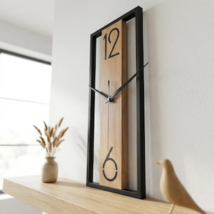Könnte beinhalten: Eine rechteckige Wanduhr mit schwarzem Rahmen und hellem Holzmaserungszifferblatt. Die Uhr hat schwarze Zeiger und große schwarze Ziffern an den Positionen 12 und 6. Die Uhr ist an einer weißen Wand befestigt.