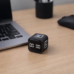 Op de afbeelding: Een zwarte kubus timer met witte digitale cijfers met de tekst "00 00", "30" en "60". De timer staat op een lichtbruin houten bureau naast een laptop en een computermuis.