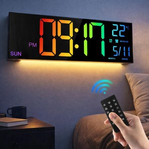 Può includere: Un orologio da parete digitale nero che mostra l'ora, la data e la temperatura. L'ora è visualizzata in grandi cifre LED colorate. Un telecomando è tenuto in mano e l'orologio è montato su una parete con luce calda.
