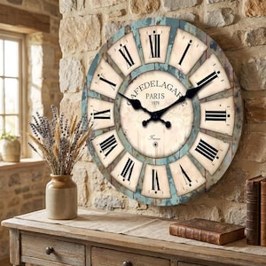 Puede incluir: Un reloj de pared grande y redondo con un diseño vintage desgastado. La esfera del reloj presenta números romanos, manecillas negras y el texto "CAFEDELAGARE PARIS 1979 France". El reloj está montado en una pared de piedra.