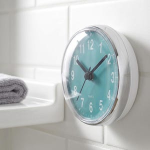 Op de afbeelding: Een ronde, witte quartz klok met een turquoise wijzerplaat en een doorzichtige plastic afdekking. De klok heeft zwarte wijzers en witte cijfers. De klok is bevestigd aan een witte betegelde muur.