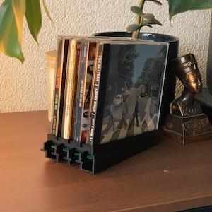 Op de afbeelding: Een zwarte cd-houder met verschillende cd's, waaronder "Abbey Road" van The Beatles. De houder staat op een houten oppervlak, met een plant en een klein beeldje op de achtergrond. De cd-hoezen zijn zichtbaar.