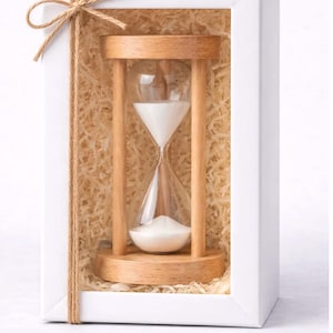 May include: An hourglass with a light-toned wooden frame and clear glass bulbs containing white sand, housed inside a white rectangular gift box filled with shredded paper filler and tied with a thin brown twine bow on the top left corner.