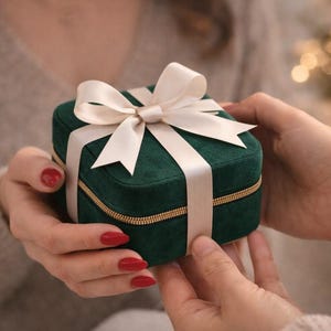 May include: A small, square, emerald green velvet gift box with a gold zip and a cream-coloured ribbon tied in a bow. The box is being held by two people, suggesting a gift exchange.