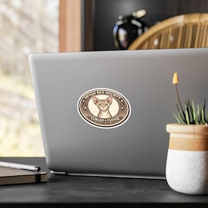 Könnte beinhalten: Ein ovaler Aufkleber mit einer Illustration einer Devon Rex Katze. Das Design enthält den Text "Devon Rex Society" und "Curled & Clever" in einem Kreis, in Hellbraun und Creme. Der Aufkleber befindet sich auf einem grauen Laptop.