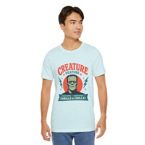 Pode incluir: Uma t-shirt azul-clara mesclada com um gráfico do monstro de Frankenstein centrado num círculo vermelho. O texto diz CREATURE FEATURE, SHOCKING! SHOCKING! e THRILLS & CHILLS! com raios azuis em ambos os lados do monstro.