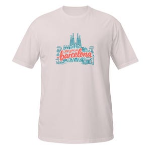 May include: A light pink t-shirt with a blue and red graphic that says "see you in barcelona". The graphic features an illustration of the Sagrada Familia and other Barcelona landmarks.