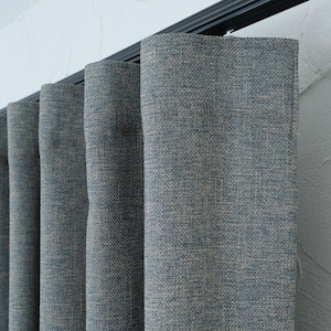 Peut inclure: Un ensemble de rideaux bleu-gris avec une texture tissée visible, suspendus à un rail noir. Le tissu présente un mélange de fils bleus et beiges, créant un aspect chiné subtil avec des plis verticaux profonds sur toute la longueur.