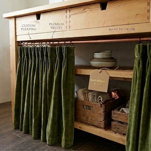 May include: Olive green velvet curtains with custom pleating hang from a copper rod on a wooden cabinet. The cabinet features drawers and open shelving containing stacked bowls, folded textiles, and wooden crates. A tag reads SerraHome Atelier.