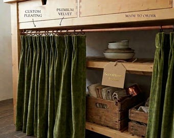 May include: Olive green velvet curtains with custom pleating hang from a copper rod on a wooden cabinet. The cabinet features drawers and open shelving containing stacked bowls, folded textiles, and wooden crates. A tag reads SerraHome Atelier.