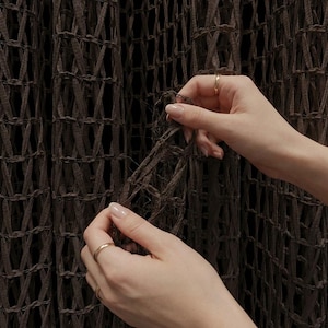 May include: Two gold bands worn on fingers, one on the ring finger and one on the index finger, against a dark brown textured mesh fabric. The rings are simple, smooth, and metallic, contrasting with the intricate, woven pattern of the dark material.
