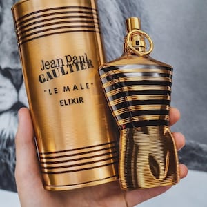 May include: A gold-toned perfume bottle and matching cylindrical container. The bottle is shaped like a torso with black horizontal stripes. The container has the text "Jean Paul Gaultier" and "LE MALE ELIXIR".