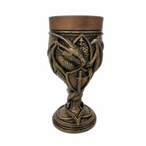 Può includere: Un calice decorativo con finitura color bronzo, caratterizzato da un drago scolpito avvolto attorno alla coppa. Il design include un emblema centrale a forma di scudo con un motivo a spada, una superficie testurizzata e un bordo superiore liscio.