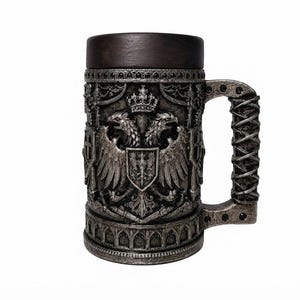 May include: A cylindrical beer stein featuring a dark brown wooden rim and a silver-toned metallic body with an embossed double-headed eagle crest topped by a crown. The handle has a textured, wrapped design, and the base displays an arched pattern.