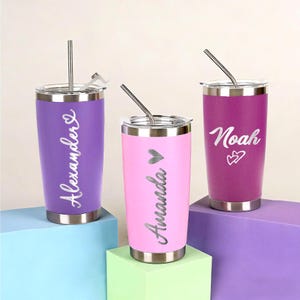Puede incluir: Tres vasos personalizados de acero inoxidable en morado, rosa y magenta. Cada vaso tiene una pajita plateada y un nombre en cursiva. El vaso rosa tiene un símbolo de corazón. Los vasos están sobre bloques coloridos.