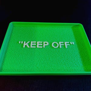 以下が含まれることがあります： 白い文字で「KEEP OFF」と書かれた、鮮やかな緑色の長方形のトレイ。トレイはテクスチャ加工された表面と盛り上がった縁があります。背景は濃い青色です。