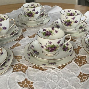 Puede incluir: Un juego de seis tazas y platillos blancos con un diseño floral púrpura y ribetes dorados. Las tazas de té están dispuestas sobre un mantel de encaje blanco. Un juego de té vintage.