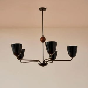 Op de afbeelding: Een moderne kroonluchter met vijf zwarte, klokvormige lampenkappen. De armatuur heeft een donker bronzen afwerking met een houten accent. Het licht hangt aan een centrale stang.
