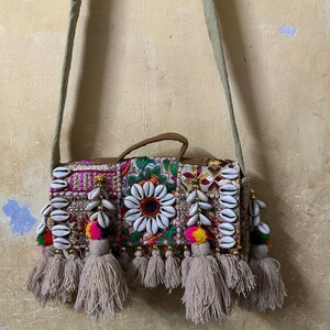 Può includere: Borsa a tracolla beige con pannello frontale decorativo. Il pannello presenta un motivo floreale con dettagli di conchiglie bianche, nappe colorate e un manico marrone. La borsa ha una tracolla lunga e regolabile.