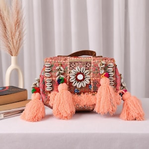 May include: A rectangular handbag featuring a pink fabric base with intricate floral embroidery, decorated with rows of white cowrie shells, a central circular mirror, and four large peach-colored yarn tassels with small multicolored pom-poms.