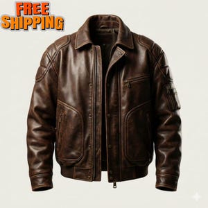 May include: A dark brown leather bomber jacket featuring a classic collar, a front zip closure with a snap-button placket, quilted shoulder panels, two side pockets with zips, and a small zipped utility pocket on the left sleeve.