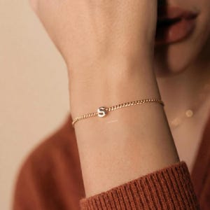 Peut inclure: Un bracelet doré avec un petit charme en forme de lettre "S" argentée. Le bracelet est une fine chaîne portée au poignet. L'arrière-plan est de ton neutre et chaud.