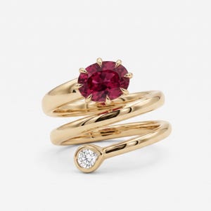 Könnte beinhalten: Ein Goldring mit polierter gelbgoldener Ringschiene, die sich spiralförmig um den Finger windet. Der Ring ist an einem Ende mit einem großen, oval geschliffenen, dunkelroten Edelstein in einer Krappenfassung und am anderen mit einem runden Diamanten besetzt.