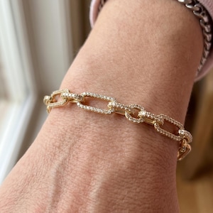 May include: A gold chain link bracelet adorned with clear, sparkling stones. The bracelet, featuring rectangular and oval links, is displayed on a wrist. This is a piece of jewellery.