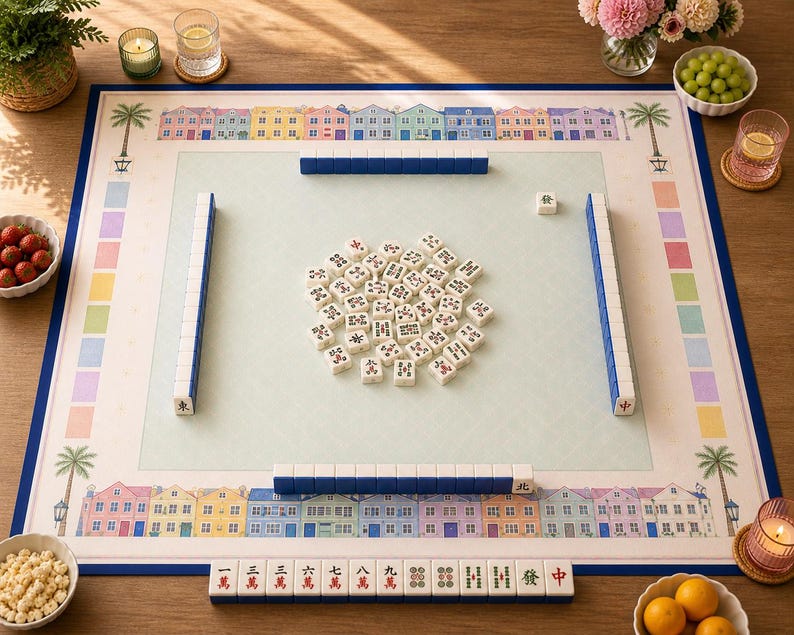 May include: A complete Mahjong set with a light blue playing surface, surrounded by a blue border. The set includes tiles, racks, and a dice. The border features colorful houses and palm trees. Various snacks and drinks are arranged around the game.