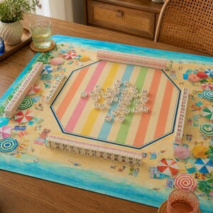 May include: A colorful mahjong set on a beach-themed mat. The mat features a sandy beach design with umbrellas and a striped center. The mahjong tiles are white with black markings, and the set includes racks.