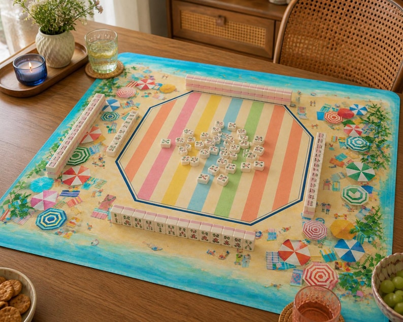 May include: A colorful mahjong set on a beach-themed mat. The mat features a sandy beach design with umbrellas and a striped center. The mahjong tiles are white with black markings, and the set includes racks.